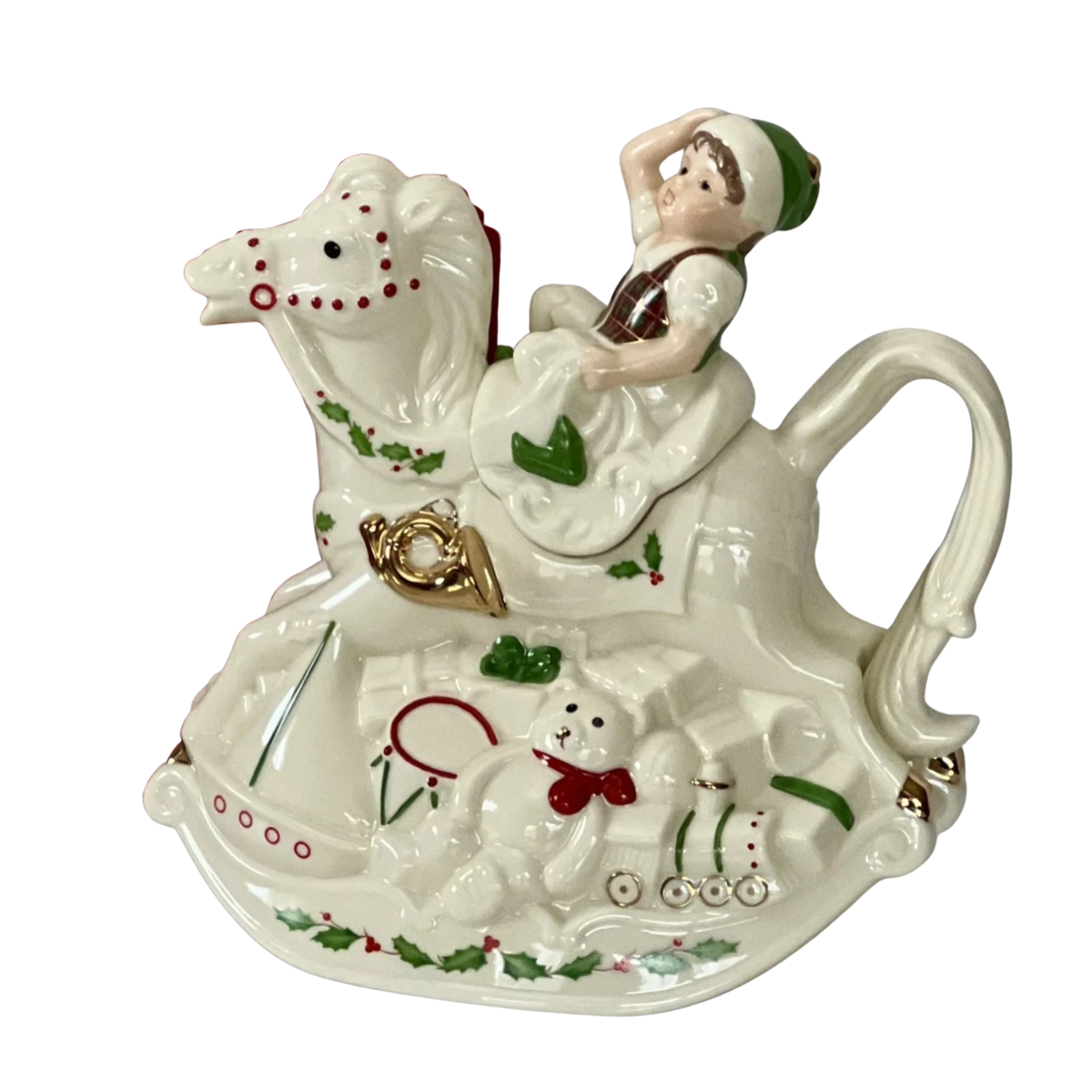 Side view of a Lenox cream colored hand-painted porcelain holiday teapot with a smiling elf in a green hat on a rocking horse with a gold horn, a teddy bear in a red bow, a drum and a train engine at the base along with a green holly design on a white background.
