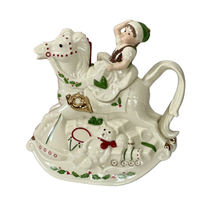 Side view of a Lenox cream colored hand-painted porcelain holiday teapot with a smiling elf in a green hat on a rocking horse with a gold horn, a teddy bear in a red bow, a drum and a train engine at the base along with a green holly design on a white background.