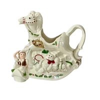 Side view of a Lenox cream colored hand-painted porcelain holiday teapot in the shape of a rocking horse and the top is a smiling elf in a green hat. The rocking horse has a teddy bear in a red bow, a drum and a train engine with green stripes and gold accents  at the base along with a green holly design on a white background. 