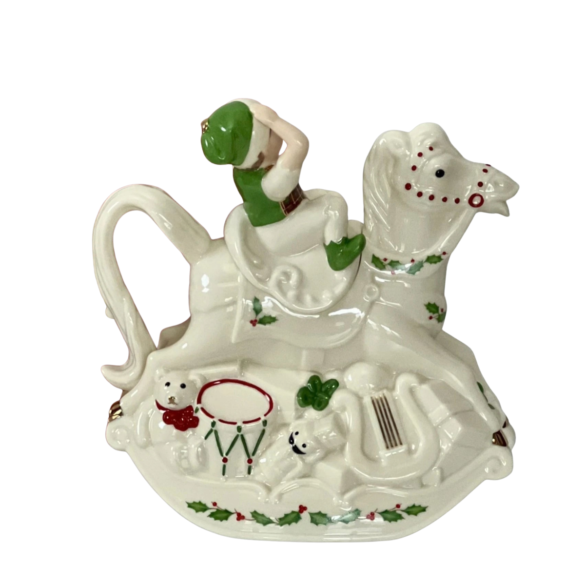 Side view of a Lenox cream colored hand-painted porcelain holiday teapot with a smiling elf in a green hat on a rocking horse with a teddy bear in a red bow, a drum and a nutcracker with a black mustache at the base on top of a green holly design on a white background. 