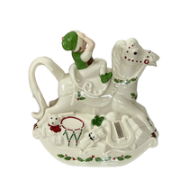 Side view of a Lenox cream colored hand-painted porcelain holiday teapot with a smiling elf in a green hat on a rocking horse with a teddy bear in a red bow, a drum and a nutcracker with a black mustache at the base on top of a green holly design on a white background. 