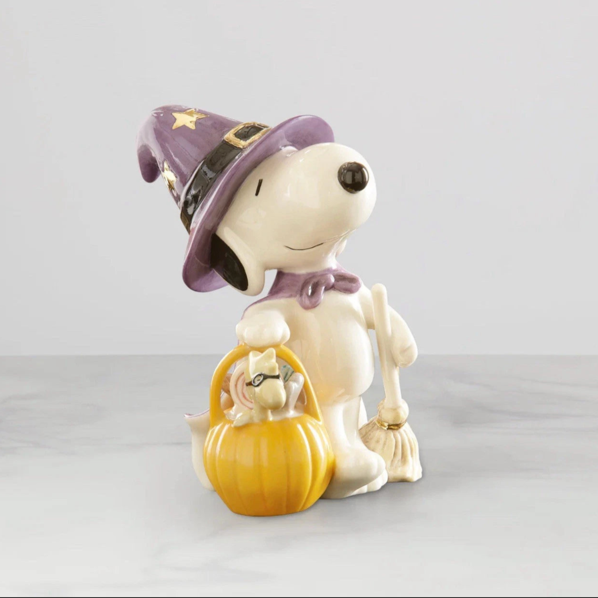 Lenox porcelain Snoopy figurine wearing a purple witch hat with a pumpkin basket holding woodstock in a black mask in one hand and a broom in the other hand on light gray background.