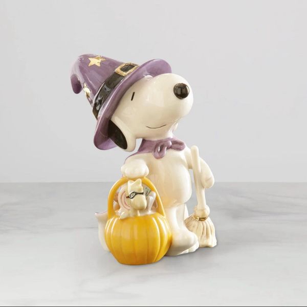 Lenox porcelain Snoopy figurine wearing a purple witch hat with a pumpkin basket holding woodstock in a black mask in one hand and a broom in the other hand on light gray background.